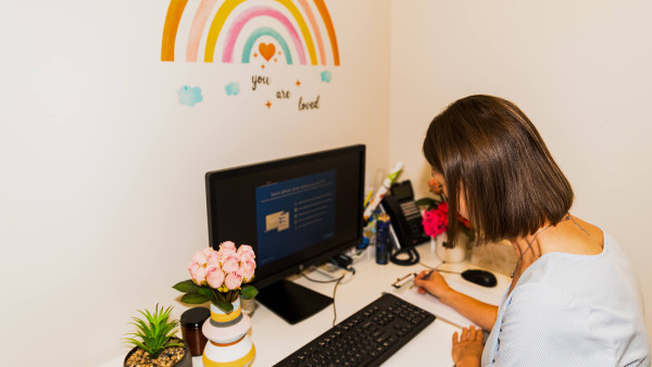 headstart person at office desk v2 A woman writes notes at a desk with a computer in a bright, welcoming office space.
